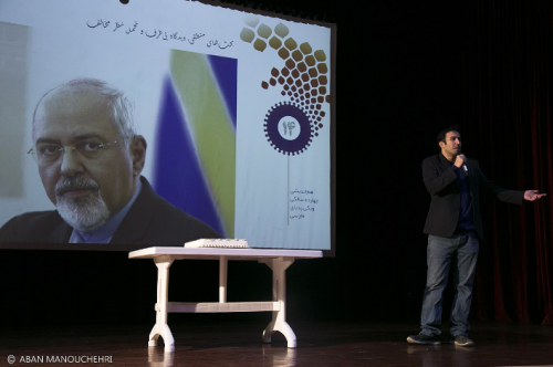 Фотография изображает человека, который выступает на сцене. Он стоит перед большим изображением Мохаммада Джавада Зарифа, министра иностранных дел Ирана. Фото: opendemocracy.net